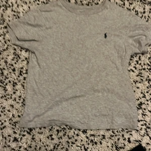 Grå t-shirt från Polo Ralph Lauren - Snygg grå t-shirt från Polo Ralph Lauren med klassisk mörkblå broderad logga på bröstet. T-shirten har rund halsringning och korta ärmar. Tillverkad i mjuk bomull som är skön mot huden. Perfekt för en clean och stilren look.