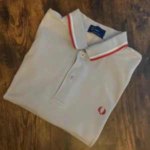 Fred Perry Pikétröja  - Fred Perry piké i ljusgrått med röda detaljer och broderad logga. Storlek Large. Klassisk twin tipped-modell med ikonisk stil. Har svettmärken men i övrigt fint skick – inga hål eller trasiga sömmar. Perfekt för dig som gillar stilen men vill fynda.