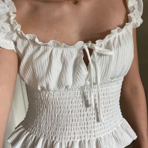 Vit blus med volang - Supersöt vit blus med smockad midja, volangkant och knytning framtill. Toppen har korta ärmar med volang. Perfekt för dig som vill ha en gullig topp i garderoben!💗Aldrig använd men har ingen prislapp kvar💗. Från Gina💗