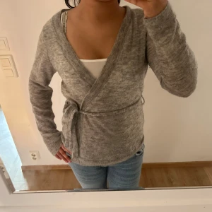 Grå omlottkofta från STOCKH LM - Mysig grå kofta från STOCKH LM i omlottmodell med knyt i midjan. Koftan är stickad och har lång ärm, perfekt för lager-på-lager och chill dagar. Den har en mjuk känsla och passar snyggt till jeans eller kjol. Stolrek M aldrig använd