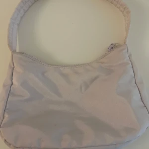Ljuslila axelväska i syntet - Säljer en stilren ljuslila axelväska i mjukt syntetmaterial. Väskan har en enkel design med vadderat handtag och dragkedja upptill. Perfekt storlek för det viktigaste och lätt att matcha med olika outfits.