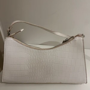 Vit axelväska med krokoprägling - Säljer en stilren vit axelväska med krokodilpräglat mönster och silverfärgad dragkedja. Väskan har en smal axelrem och avlång form, perfekt för att bära det viktigaste. Materialet är syntet och ytan har en lyxig känsla.