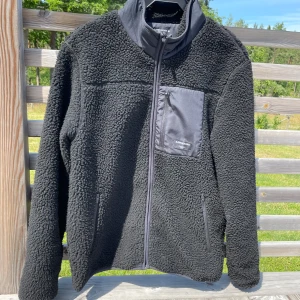 Svart teddyjacka från Björn Borg S - Svart teddyjacka från Björn Borg i storlek S. Jackan har mjuk och fluffig fleece, hög krage och dragkedja framtill. Praktisk bröstficka med dragkedja och två sidofickor. Perfekt för kyliga dagar när du vill vara både bekväm och snygg.