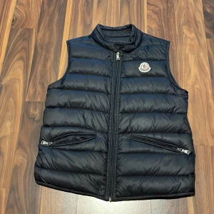 Svart dunväst från Moncler - Snygg svart dunväst från Moncler med klassisk quiltad design och hög krage. Västen har två fickor med dragkedja och Moncler-logga på bröstet. Perfekt att slänga över en hoodie för en cool look.