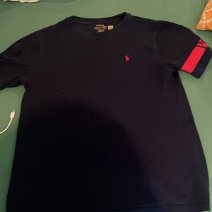 Mörkblå Polo Ralph Lauren t-shirt - Snygg mörkblå t-shirt från Polo Ralph Lauren med klassisk röd broderad logga på bröstet och en röd detalj på ena ärmen. T-shirten har rund hals och är tillverkad i mjuk bomull. Perfekt för en clean och stilren look.