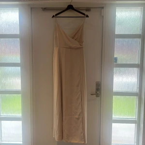 Champagnefärgad långklänning med omlott - Elegant långklänning i champagnefärg med tunna axelband och omlottdesign framtill. Klänningen har en snygg knytning i ryggen och är tillverkad i ett glansigt, mjukt material som ger en lyxig känsla. Använd en gång. 