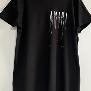 Svart Amiri t-shirt med färgstänk - Svart t-shirt från Amiri med coolt tryck framtill och baktill. Stora vita bokstäver och färgstänk i blått, vitt, rött och orange som rinner ner över tyget. Klassisk passform och rund hals. Perfekt för dig som gillar streetstyle och statement-plagg.