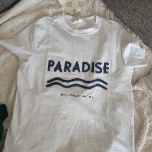 Vit t-shirt med blått PARADISE-tryck prislapp kvar - Vit t-shirt i bomull med blått glittrigt PARADISE-tryck och vågmotiv på bröstet. Texten 'BEACH RESORT, ANTIBES' under motivet. Klassisk rund hals och korta ärmar. Perfekt för en chill sommarlook. Med prislapp. Slutsåld på hm.com nu. 