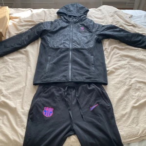 FC Barcelona Tracksuit - Svart FC Barcelona vindjacka från Nike med huva och hel dragkedja. Jackan har klubbmärke och Nike-logga i lila och rosa på bröstet samt diskret mönstrad ovandel. Perfekt för dig som älskar fotboll och vill ha en sportig look.