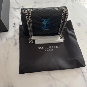 axelväska från Saint Laurent - Säljer en svart quiltad axelväska från Saint Laurent med silverfärgad kedjerem och ikoniskt YSL-emblem framtill. Väskan är tillverkad i skinn och har ett stilrent, lyxigt uttryck med dragkedjefack inuti. Perfekt för dig som vill ha en klassisk och trendig väska.