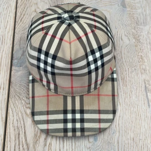 Burberry rutig keps i beige och svart - Snygg keps från Burberry med rutigt mönster i beige, svart, vitt och röda linjer. Justerbar baktill med svart spänne och tydlig Burberry.