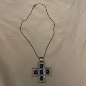 Halsband med blått korshänge - Unikt halsband med ett stort korsformat hänge i silverfärg. Hänget är dekorerat med sju fyrkantiga, djupblå stenar och gnistrande små stenar runtom. Kedjan är tunn och enkel, vilket gör att det maffiga hänget verkligen står ut. Perfekt för dig som gillar statement-smycken.