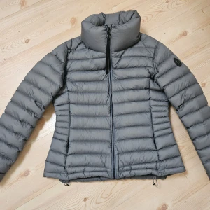 Grön/grå dunjacka från 8848 Altitude stl M/L - Snygg grön/grå dunjacka från 8848 Altitude med hög krage och horisontella sömmar. Jackan har dragkedja framtill och justerbar nederkant. Perfekt för kalla dagar när du vill hålla stilen och värmen. Storlek 44 men mer som 42/44 alltså M/L.