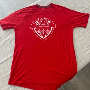Röd Under Armour t-shirt Kohl's - Röd t-shirt från Under Armour med vit tryckt logga och motiv från Kohl's National Scholarship Camp på bröstet. Klassisk rund hals, korta ärmar och loose fit. Perfekt för dig som gillar sportig stil och vill sticka ut med en bold färg.