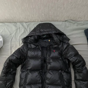 Svart dunjacka Polo Ralph Lauren - Svart dunjacka från Polo Ralph Lauren med avtagbar huva och klassisk röd logga på bröstet. Jackan har dragkedja framtill och två sidofickor med dragkedja. Perfekt för kalla dagar och riktigt snygg streetstil.