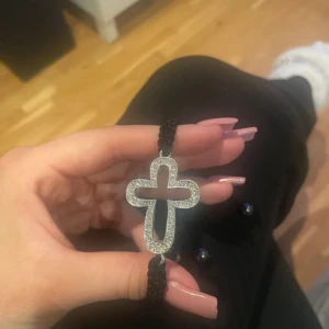 Svart armband med glittrigt kors - Snyggt svart armband med flätad snodd och ett stort kors i silverfärg, täckt av glittrande stenar. Knytband med små pärlor i ändarna gör det enkelt att justera storleken. Perfekt accessoar för dig som gillar statement-smycken och bling.