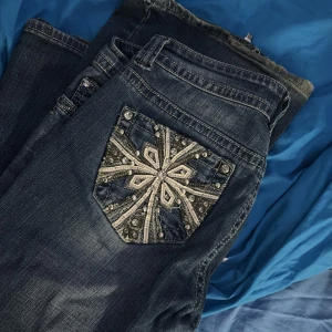 Premiere jeans med blingad bakficka - Säljer ett par blå jeans från Premiere med en riktigt snygg bakficka dekorerad med pärlor, paljetter och broderier i ett stjärnmönster. Jeansen har klassisk femficksdesign, silverfärgade detaljer och normal passform. Perfekta för dig som gillar att sticka ut lite extra!