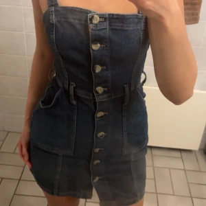 Mörkblå jeansklänning med bälte - Snygg mörkblå jeansklänning med knappar hela vägen framtill och tillhörande bälte i midjan. Klänningen är ärmlös med breda axelband och har praktiska fickor på sidorna. Perfekt för dig som gillar en cool och trendig denimlook.