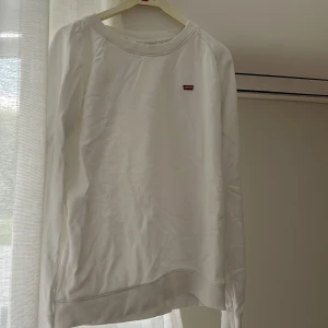 Vit sweatshirt från Levi's - Klassisk vit sweatshirt från Levi's med liten röd logotyp på bröstet. Tröjan har rund halsringning och långa ärmar, perfekt för en clean och enkel stil. Tillverkad i mjuk bomull som känns skön mot huden.