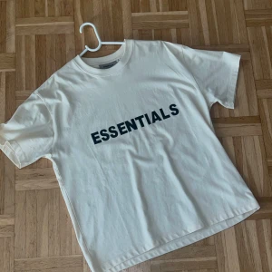 Vit Essentials Fear of God t-shirt - Vit t-shirt från Essentials Fear of God med svart tryck på bröstet. Klassisk rund hals och korta ärmar. Oversized fit för en avslappnad vibe. Tillverkad i mjuk bomull som känns skön mot huden. Perfekt för dig som gillar clean och stilren streetwear.