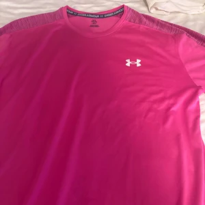 Rosa tränings-tshirt från Under Armour - Snygg rosa t-shirt från Under Armour med korta ärmar och rund hals. Tillverkad i ett lätt och snabbtorkande material som passar perfekt för träning. Diskret logga på bröstet och coola detaljer på axlarna. Den här är andvänd 1 gång.