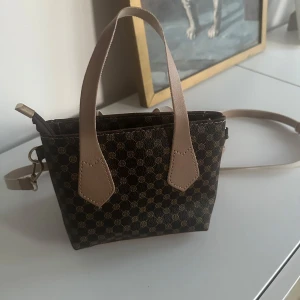 Liten Brun mönstrad handväska med axelrem - Snygg brun handväska med ett klassiskt mönster och ljusa handtag. Väskan har en avtagbar axelrem och dragkedja upptill. Perfekt för att bära dina nödvändigheter med stil. Knappt använd 