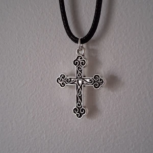 Silverfärgat korshalsband - Elegant halsband med ett detaljerat korshänge i silverfärg. Hänget har vackra snirkliga mönster och hänger på en svart rem. Perfekt för att ge en stilfull touch till din outfit.