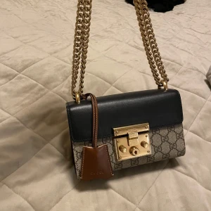 Axelväska från Gucci - Snygg axelväska från Gucci med klassiskt mönster och guldfärgad kedja. Väskan har en elegant låsdetalj i guld och en kontrasterande brun läderdetalj. Perfekt för att ge en lyxig touch till din outfit.