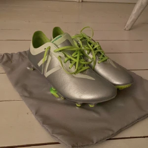 Silvriga fotbollsskor från New Balance  - Snygga fotbollsskor i silver med gröna detaljer från New Balance. Skorna har snörning och en blänkande sula som ger ett futuristiskt intryck. Perfekta för den som vill sticka ut på planen.