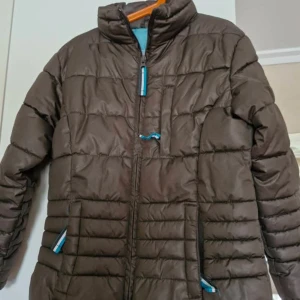 Brun dunjacka från Covalliero - Säljer en brun dunjacka från Covalliero med dragkedja och hög krage. Jackan har en quiltad design och är perfekt för kyligare dagar. Den har långa ärmar och en snygg logga på baksidan. Perfekt för dig som gillar en stilren look med en touch av equestrian style.