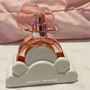 Parfym från Ariana Grande - Charmig parfymflaska med rosa vätska och ett hjärtformat lock. Flaskan står i en vit molnformad bas med märket Ariana Grande tryckt på. Perfekt för den som älskar söta och lekfulla designinslag.