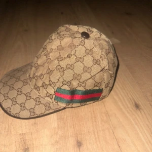 Beige keps med monogrammönster - Snygg beige keps med ett klassiskt monogrammönster och en grön och röd randdetalj på sidan. Perfekt för att ge en stilren touch till din outfit.