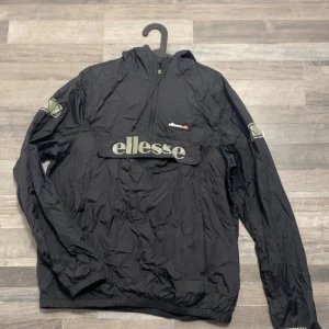 Svart vindjacka från Ellesse - Säljer en svart vindjacka från Ellesse med halv dragkedja och stor logga på bröstet. Jackan har en huva och elastiska muddar vid ärmslut och nederkant. Perfekt för blåsiga dagar. På jackan står det att det är storlek XL (vilket jag tror är ett produktionsfel) för att den passar mina vänner som har storlek S-M i jackor och liknande. Det skrynkliga går, det är bara att tvätta den. Levererar samma dag som beställningen. Priset kan diskuteras vid snabb och smidig affär.