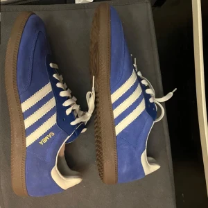 Blå Adidas Samba sneakers - Snygga blå Adidas Samba sneakers med vita detaljer och klassiska tre ränder på sidan. Skorna har en brun sula och är tillverkade i mocka. Perfekta för en sportig look. Storlek 43,3! Skriv till mig om du har frågor eller prisförslag. De är endast använda en gång, märks inte. Annars nya.  Lite skrynkliga på insidan men inget som märks när den är på!