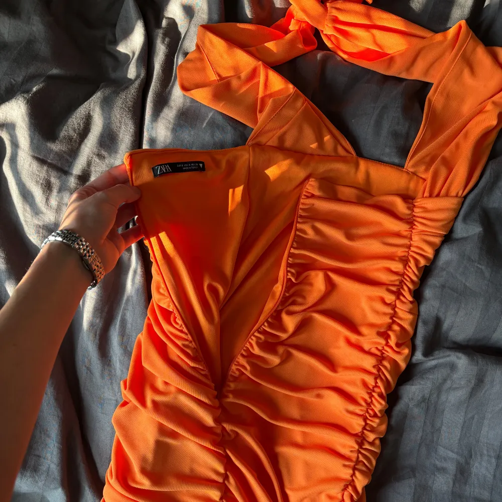 Orange omlottklänning med halterneck och rynkade detaljer. Går att knyta på 2 olika sätt (jmfr bild 1-2) öppen rygg och figurnära. Aldrig använd och normal i storleken.. Mekot.