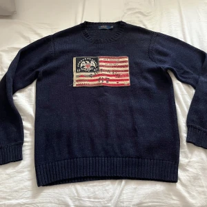 Mörkblå tröja från Polo Ralph Lauren - Säljer en snygg mörkblå tröja från Polo Ralph Lauren med ett unikt tryck på framsidan. Tröjan har långa ärmar och ribbade muddar vid ärmslut och nederkant. Perfekt för en stilren look.
