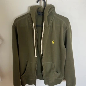 Olivgrön hoodie från Ralph Lauren - Säljer en snygg olivgrön hoodie från Ralph Lauren med dragkedja och två fickor framtill. Den har en klassisk design med en gul logotyp på bröstet och vita snören i huvan. Perfekt för en avslappnad stil.