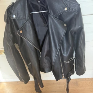 Svart skinnjacka  - Snygg svart skinnjacka med dragkedja och knappar. Jackan har en klassisk bikerstil med bälte i midjan och långa ärmar. Perfekt för våren! Använd men i väldigt bra skick, inga defekter!