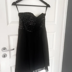 Svart axelbandslös klänning från Vila - Snygg svart axelbandslös klänning från Vila med glittrig överdel. Perfekt för en festkväll! Klänningen har en kort längd och en elegant design som verkligen sticker ut.