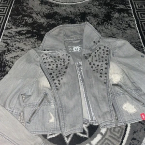 Grå jeansjacka med nitar - Säljer en grå jeansjacka från Ecko med coola nitar på axlarna och slitna detaljer. XL men passar till M. Jackan har dragkedja framtill och två dragkedjefickor. Perfekt för en edgy look!
