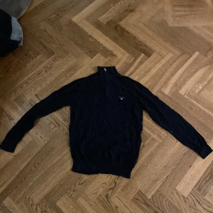 Svart tröja med dragkedja från Gant - Säljer en stilren svart tröja från Gant med dragkedja vid halsen. Tröjan har långa ärmar och en liten logotyp på bröstet. Perfekt för en avslappnad och snygg look.