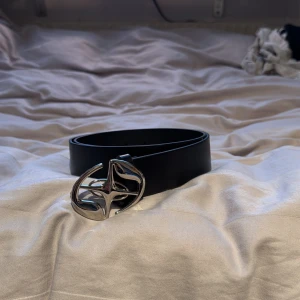Svart bälte med silverfärgat spänne - Snyggt svart bälte med ett unikt silverfärgat spänne. Perfekt för att ge din outfit en stilren touch. Passar bra till jeans och andra byxor.