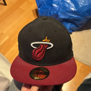 Svart och vinröd keps med NBA-logga New era - Snygg keps från New Era med svart kulle och vinröd skärm. Framsidan pryds av ett broderat emblem och baksidan har en NBA-logga. Perfekt för sportfantaster! (57,7cm)