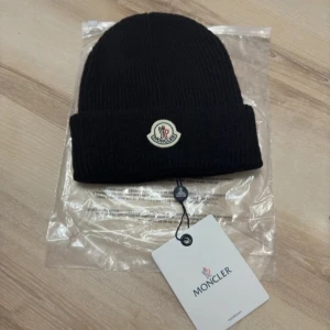 Svart ribbad mössa från Moncler - Snygg svart ribbad mössa från Moncler med deras ikoniska logga framtill. Perfekt för att hålla värmen under kyliga dagar. Stilren design som passar till det mesta.