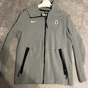 Nike tech Chelsea FC - Säljer en grå träningsjacka från Nike med Chelsea FC-logga på bröstet. Jackan har dragkedja framtill och två svarta dragkedje fickor.