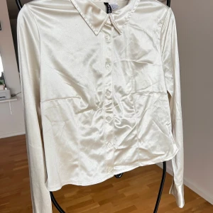 Vit/beige satinblus med knappar - Elegant vit blus i satin med långa ärmar och knappar framtill. Perfekt för en stilren look. Har lite skavanker enligt bild 2. 95% polyester, 5% elastan. 