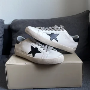 Golden Goose Superstar - Snygga vita sneakers från Golden Goose med en distinkt svart stjärna på sidan. Skorna har svarta detaljer på hälen och grå snörning. Perfekta för en stilren look. Endast använda ett fåtal gånger inomhus samt 1 gång utomhus. Priset kan förhandlas vid snabb affär.
