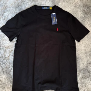 Svart t-shirt från Polo Ralph Lauren - Säljer en klassisk svart t-shirt från Polo Ralph Lauren med det ikoniska röda broderade logotypen på bröstet. T-shirten är kortärmad och tillverkad i mjuk bomull. Perfekt för en stilren look.