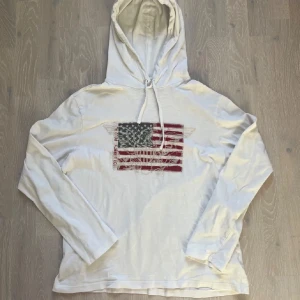 Vit hoodie från Polo Ralph Lauren - Säljer en vit hoodie från Polo Ralph Lauren med ett amerikanskt flaggapå bröstet. Tröjan har långa ärmar och en justerbar huva med dragsko. Perfekt för en avslappnad stil.