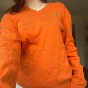 Orange tröja från Ralph Lauren - Säljer en snygg orange tröja från Ralph Lauren med v-ringning och långärmade. Den har en liten grön logga på bröstet och ribbade muddar vid ärmslut och nederkant.